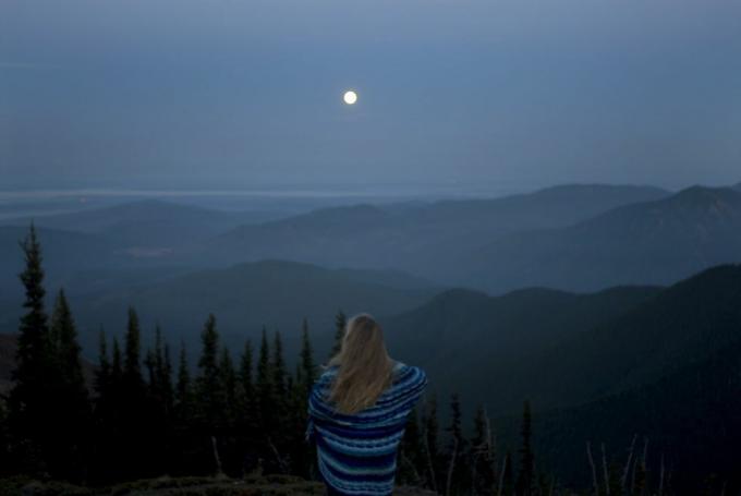 donna avvolta in una coperta che guarda il paesaggio montuoso con la luna piena, vista posteriore