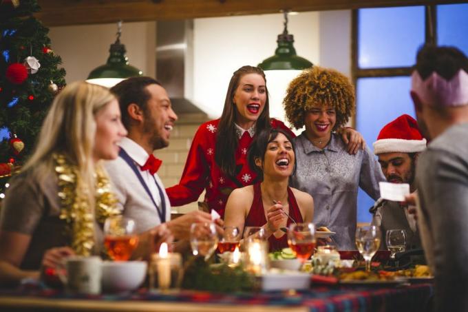 gruppo di amici che ridono mentre si siedono attorno a un tavolo mangiando e bevendo a Natale un uomo sta leggendo una barzelletta da un cracker di Natale si indossano cappelli da festa e maglioni di Natale