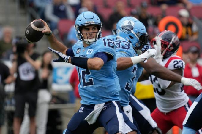 quarterback di calcio canadese in uniforme blu che lancia un passaggio mentre la linea offensiva tiene a bada un difensore in bianco e rosso