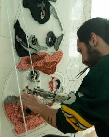Uomo barbuto ispano-latino di 30 anni che indossa una maglia da rugby, nel suo laboratorio lavora su tappeti fatti a mano con canne tufting e fibre acriliche, con un disegno di tappeto per cani