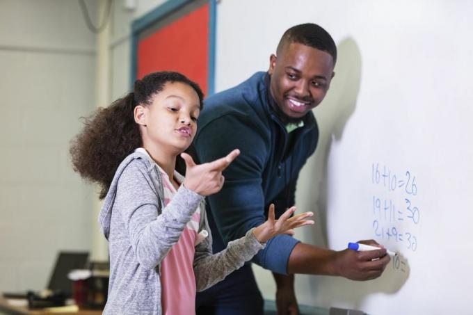 una bambina multirazziale di 9 anni a lezione di matematica, in piedi alla lavagna con la sua insegnante, un giovane afroamericano nel suo 20 anni l'insegnante guarda la sua studentessa delle elementari, aspettando mentre cerca di capire la risposta su cui si concentra l'attenzione ragazza