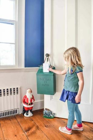 una bambina con i capelli biondi che indossa una camicetta a righe blu e verdi e dei collant sta facendo scorrere un foglio in una cassetta delle lettere verde che è appeso a una porta all